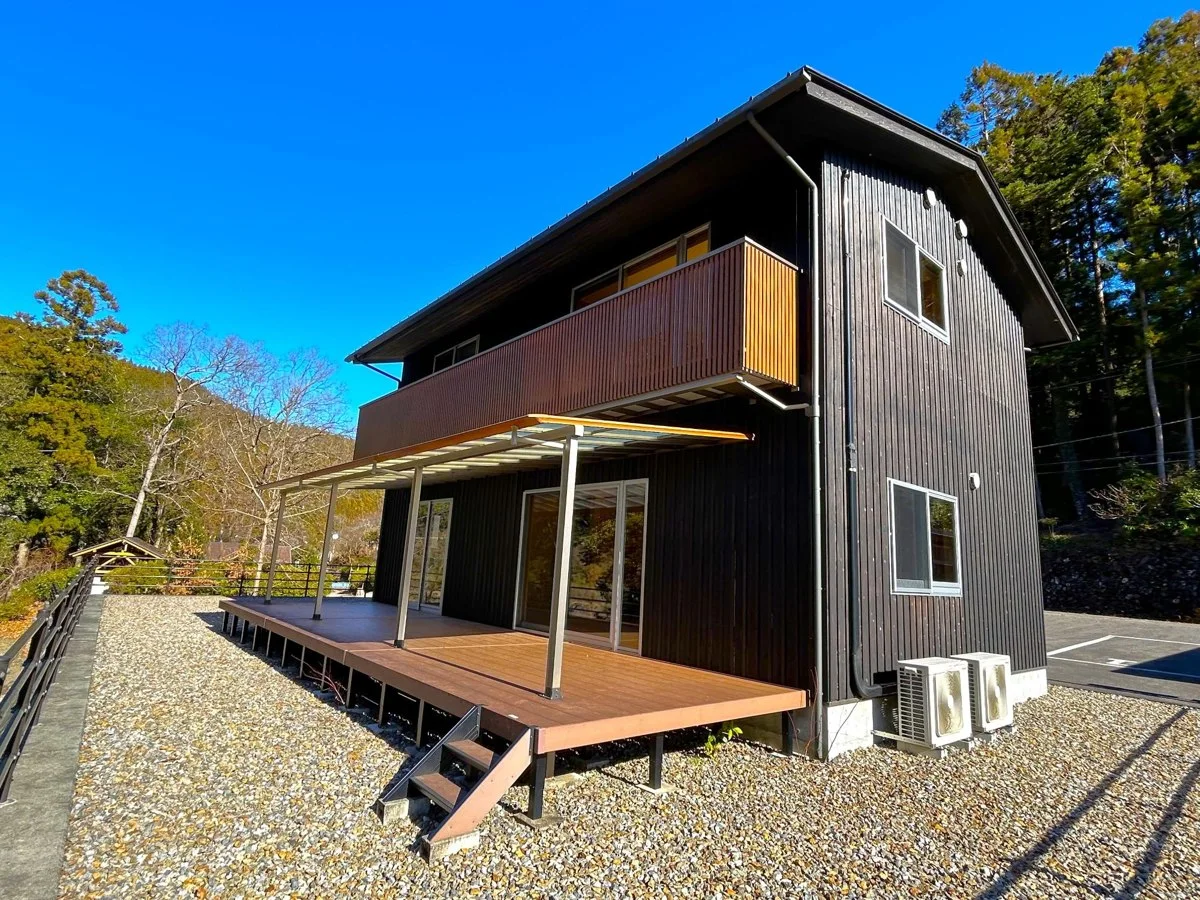 わじきライン林間キャンプ村：鉄製の柵で区切られた敷地内でベランダとウッドデッキのある黒い外観のコテージの写真