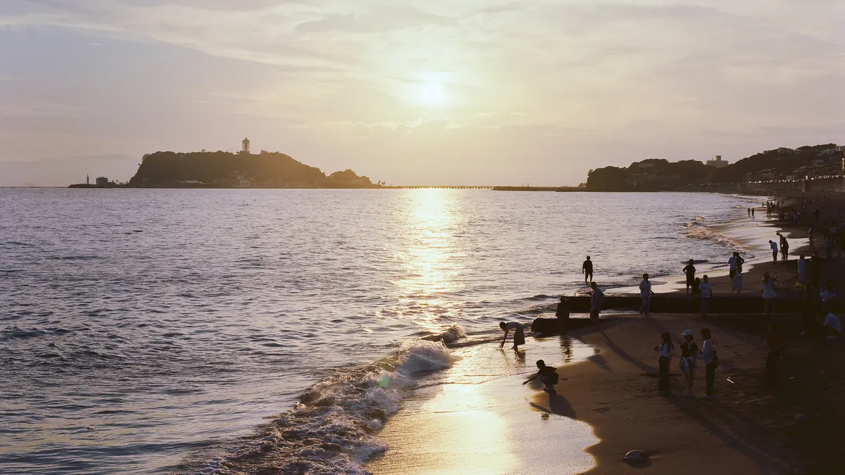 湘南の砂浜から、夕日に照らされた江ノ島と海を望んでいる(イメージ)